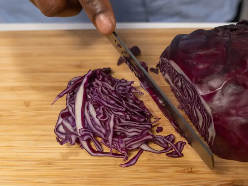 Hände schneiden frischen Rotkohl auf einem Holzbrett, daneben eine Möhre.