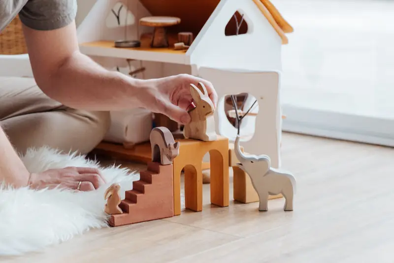 Die Hand einer Erwachsenen stellt Holztiere an ein weißes Puppenhaus aus Holz.